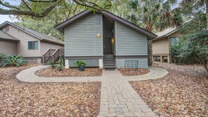 A two-story building with a covered porch, a brick pathway leading to the entrance, and a small garden area with a palm tree and potted plants. - Kiawah Resort snowbird hideaway with AC, steps to beach, near golf & tennis (Kiawah Island)