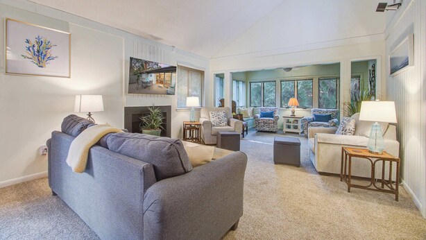A spacious living room with a grey sofa, a coffee table, and a small side table with a lamp. There is a fireplace, a bookshelf, and a view of the outdoors through large windows. - Kiawah Resort snowbird hideaway with AC, steps to beach, near golf & tennis (Kiawah Island)