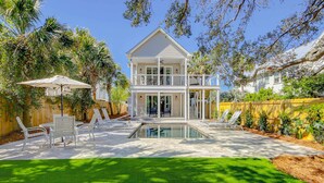 A two-story house with a pool, a patio with chairs and an umbrella, and a well-maintained lawn. - Gorgeous home with private pool, deck, enclosed patio, central AC, & balcony (Isle of Palms)