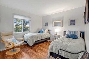 A bedroom with two beds, a chair, a bedside table with a lamp, and a window with a view of trees.