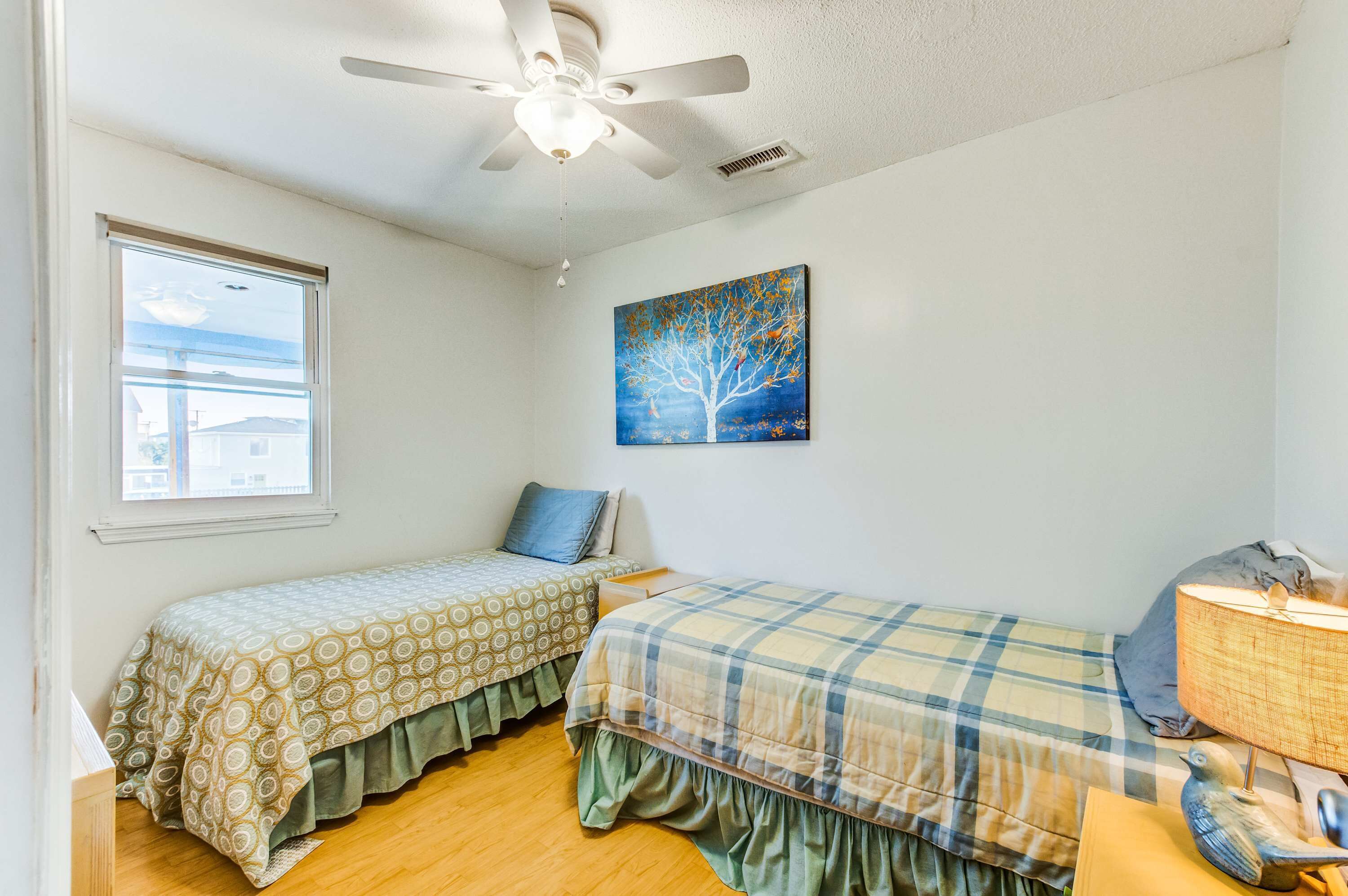 A bedroom with two single beds, a ceiling fan, a window, and a painting on the wall.