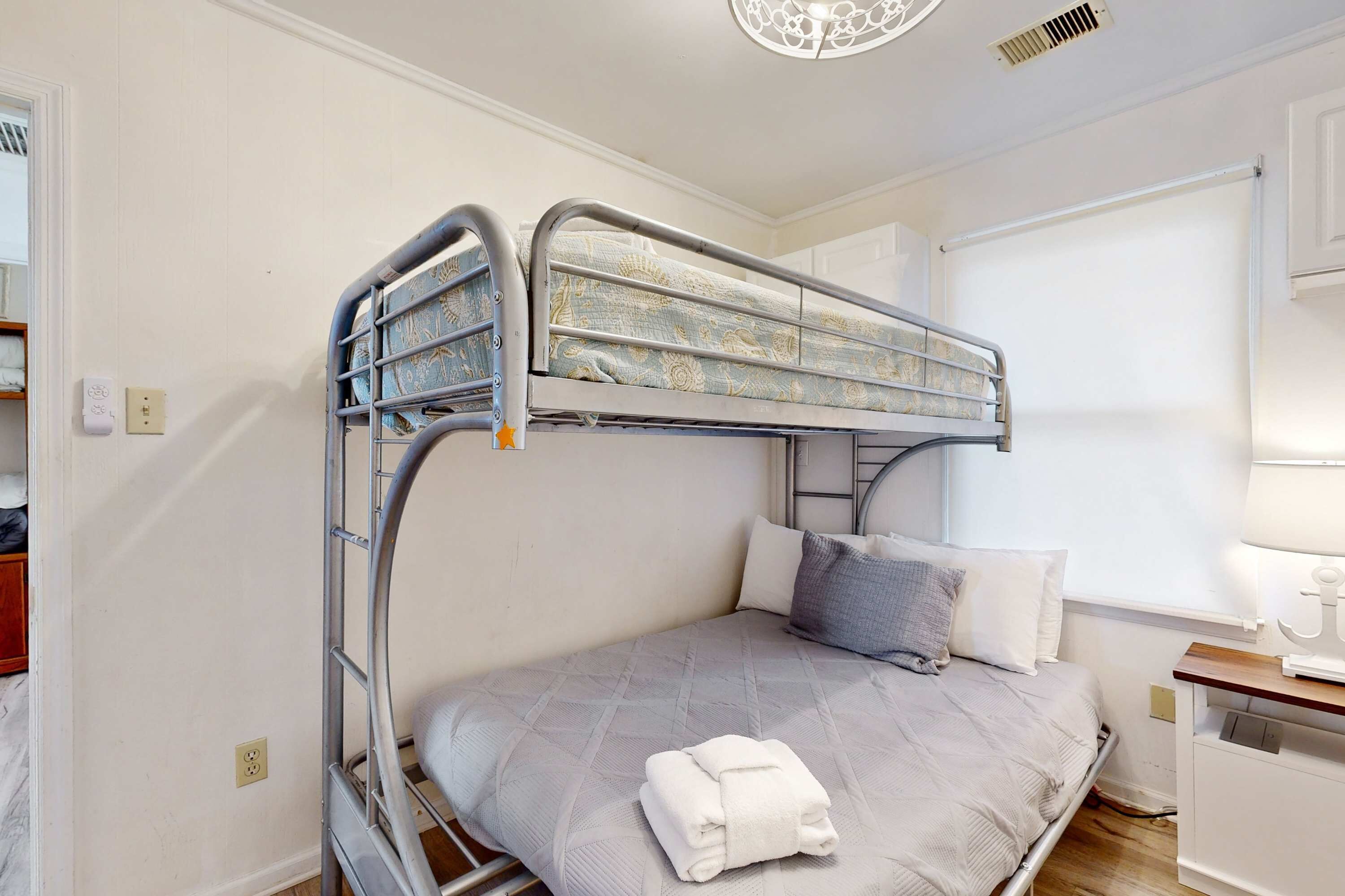A bunk bed with a grey mattress and pillows, a small bedside table with a lamp, and a window with blinds.