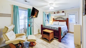 A bedroom with a bed, a television mounted on a stand, a chair, a small table, and a ceiling fan. - Beach Across the Street Ocean View Home w/ Roof Deck, Porch, AC - Dogs OK (Folly Beach)