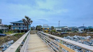 A wooden boardwalk leading to a beachfront property with multiple buildings, including a blue house with a balcony and a white house with a porch. - Beach Across the Street Ocean View Home w/ Roof Deck, Porch, AC - Dogs OK (Folly Beach)
