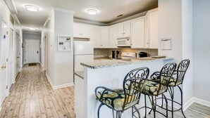 A kitchen with white cabinets, a granite countertop, and a microwave. There are two bar stools with floral upholstery. - Fantastic condo with water-view, pool, balcony, central AC, & washer/dryer (Folly Beach)
