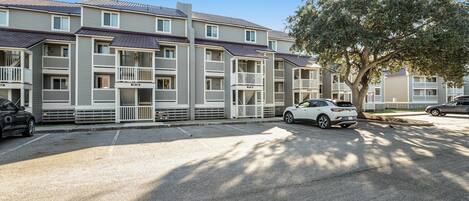 Un edificio residencial de varios pisos con balcones, un estacionamiento y un árbol en primer plano.