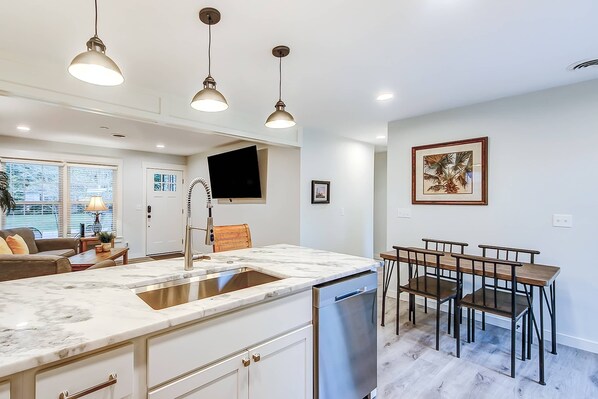 A modern kitchen with a marble countertop, stainless steel sink, and a dishwasher. There is a dining area with a table and chairs.