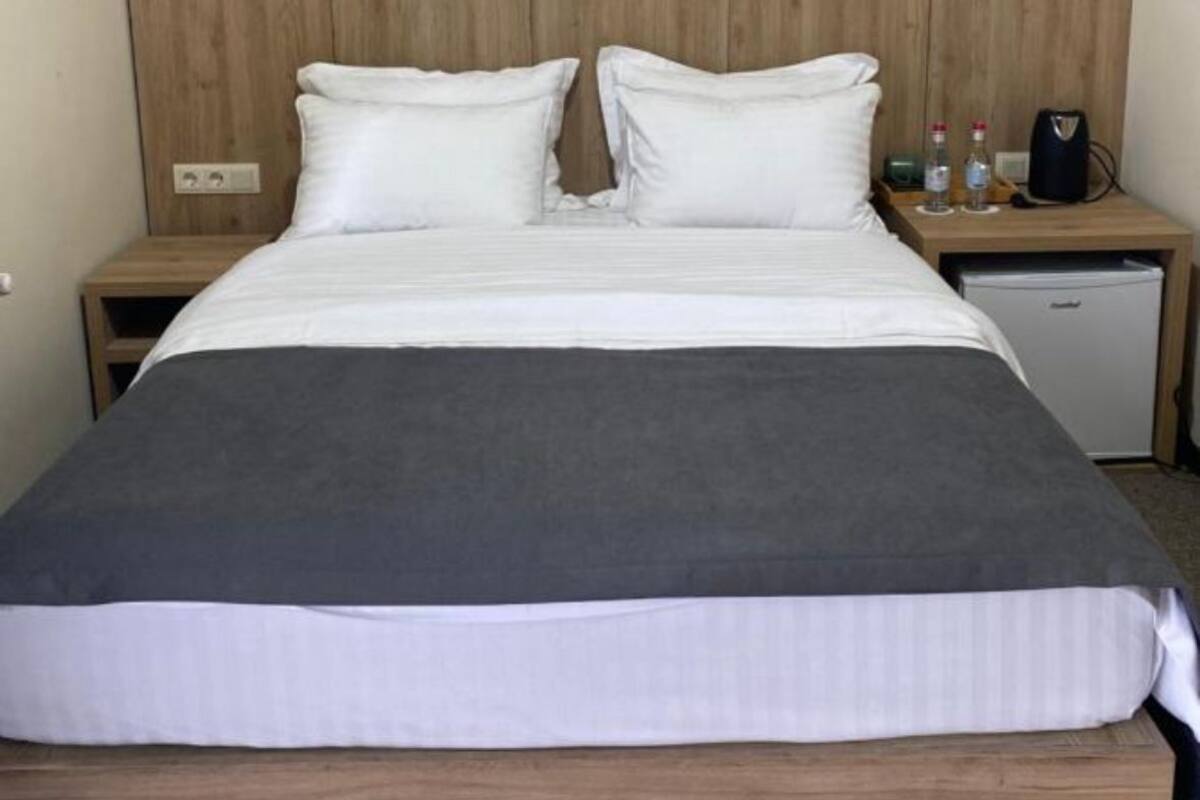 A single bed with a wooden headboard, white bedding, and a small bedside table with a lamp and a bottle.
