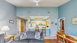 A living room with a sofa, a dining table with chairs, a kitchen in the background, and a door leading to another room.