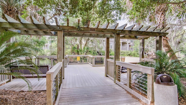 A wooden deck with a pergola, surrounded by lush greenery and a view of a building in the background.