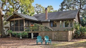 Une maison en bois avec une cheminée, de grandes fenêtres et deux fauteuils Adirondack verts dans le jardin.