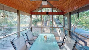 Une terrasse couverte avec une table et des chaises, un ventilateur de plafond et de grandes fenêtres donnant sur l’extérieur.