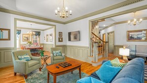 A spacious living room with a wooden coffee table, a blue sofa, and a green armchair. A staircase leads to an upper level, and a chandelier hangs from the ceiling. - Picturesque and Private near Massanutten, Sleeps 20 (Elkton)