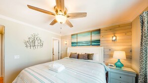 Une chambre à coucher avec un lit, un ventilateur de plafond, une table de chevet avec une lampe et un mur orné d’un tableau.