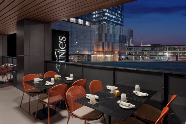 A rooftop dining area with tables set for four, orange chairs, and a city view at dusk. - Motto by Hilton Nashville Downtown (Nashville)