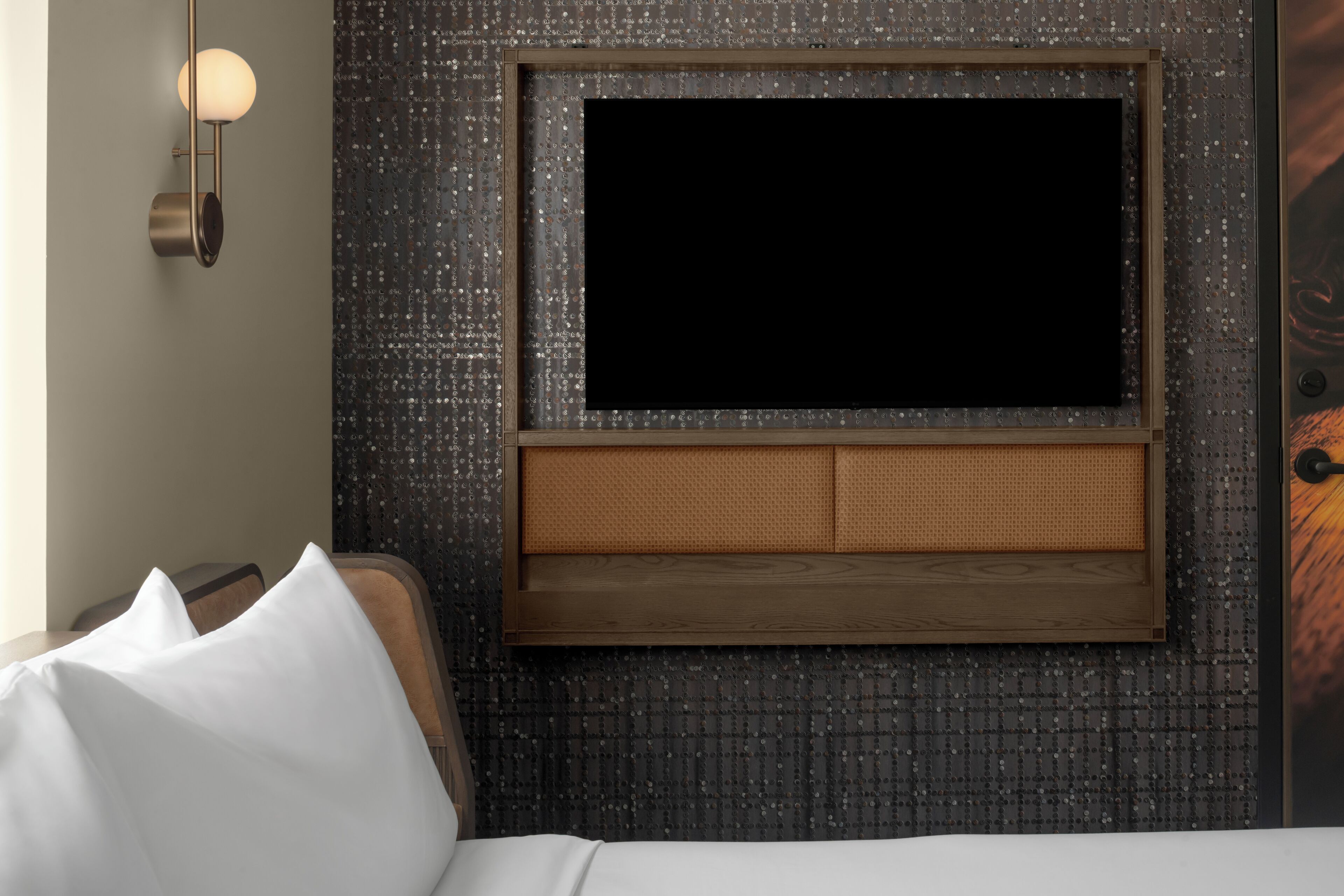 A modern hotel room with a large flat-screen TV mounted on a textured wall, a wooden headboard, and a wall-mounted light fixture.