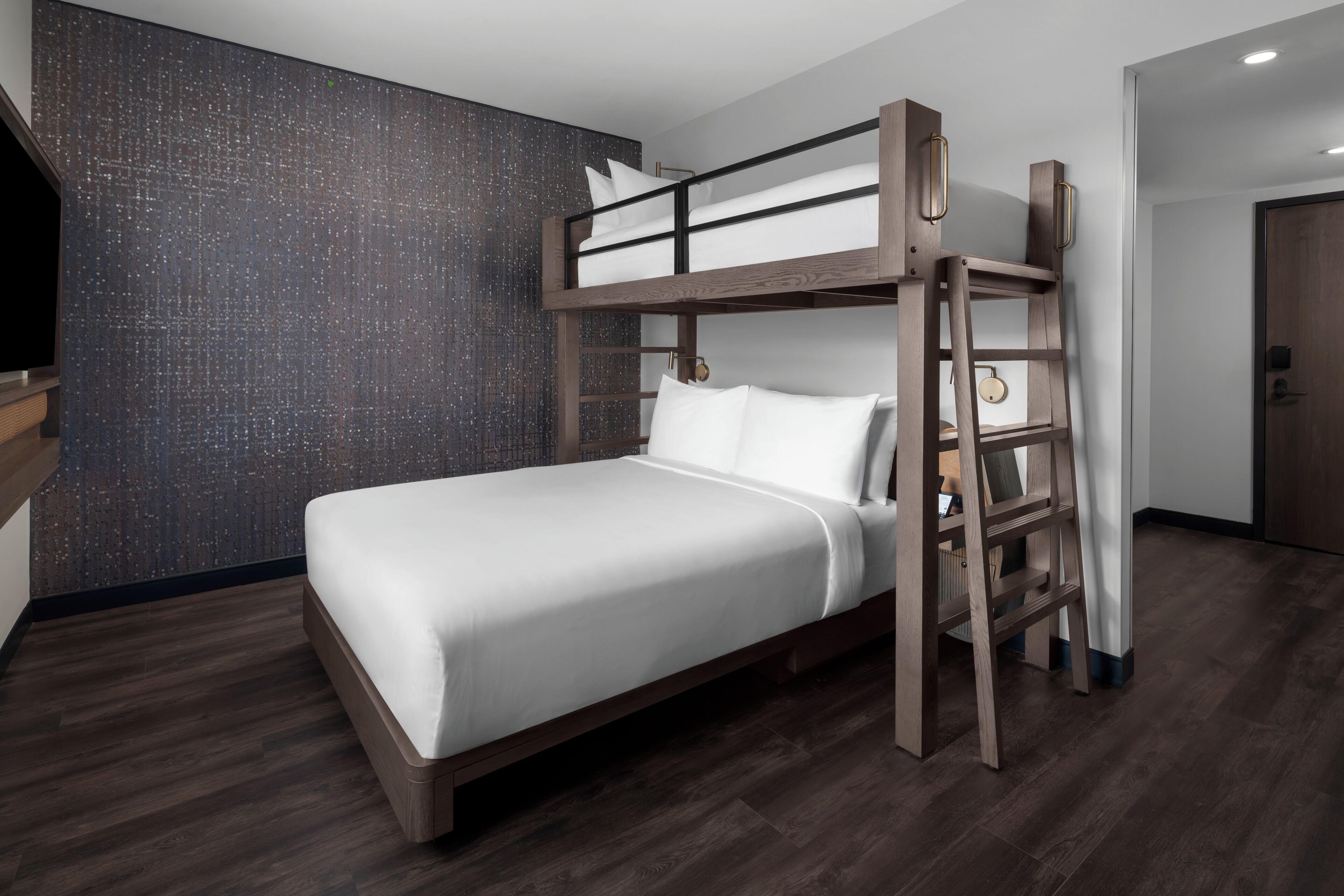 Una cama litera con estructura de madera y ropa de cama blanca en una habitación moderna con suelo de madera oscura y pared texturizada.