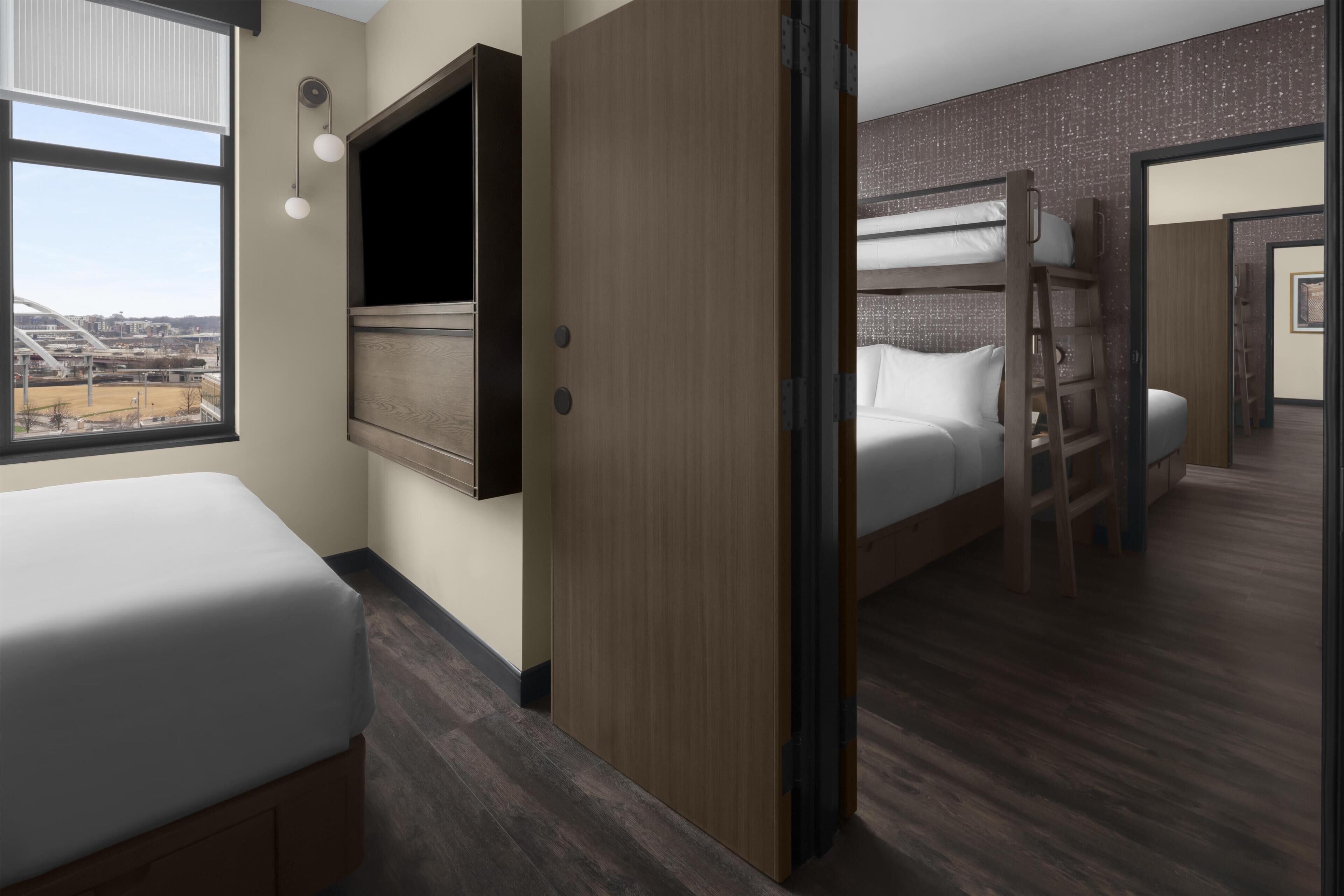 Une chambre d’hôtel avec un lit, une armoire, une fenêtre donnant sur la ville et un lit superposé dans la chambre adjacente.