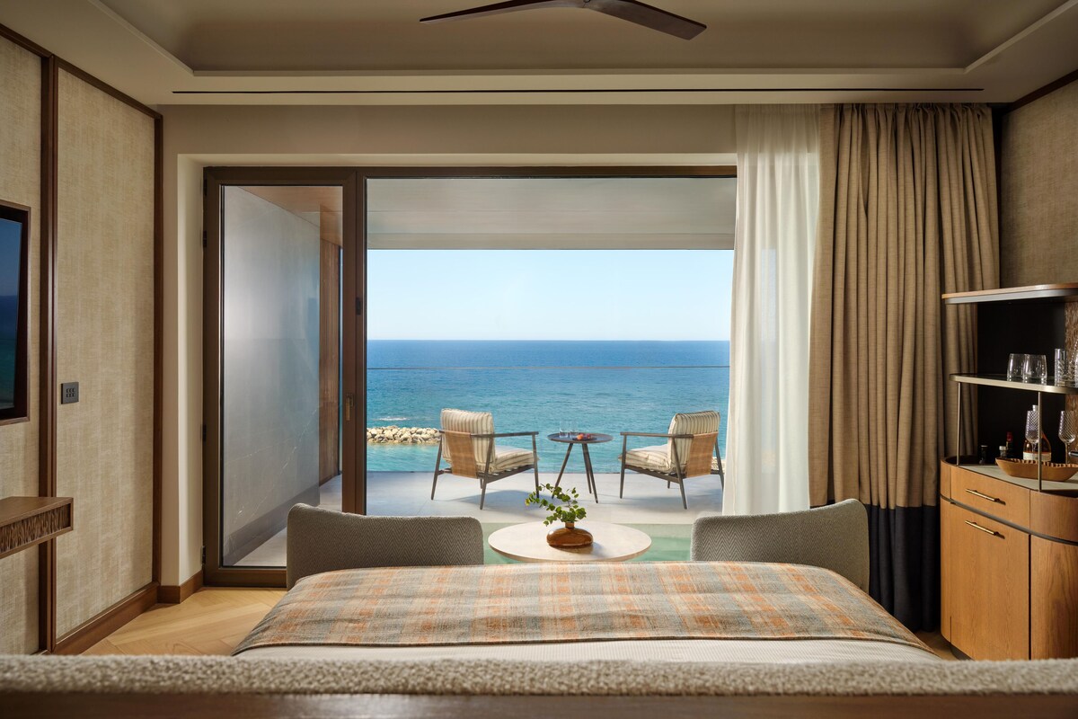 A bedroom with a bed, a ceiling fan, a view of the ocean, a small table with chairs, and a minibar.