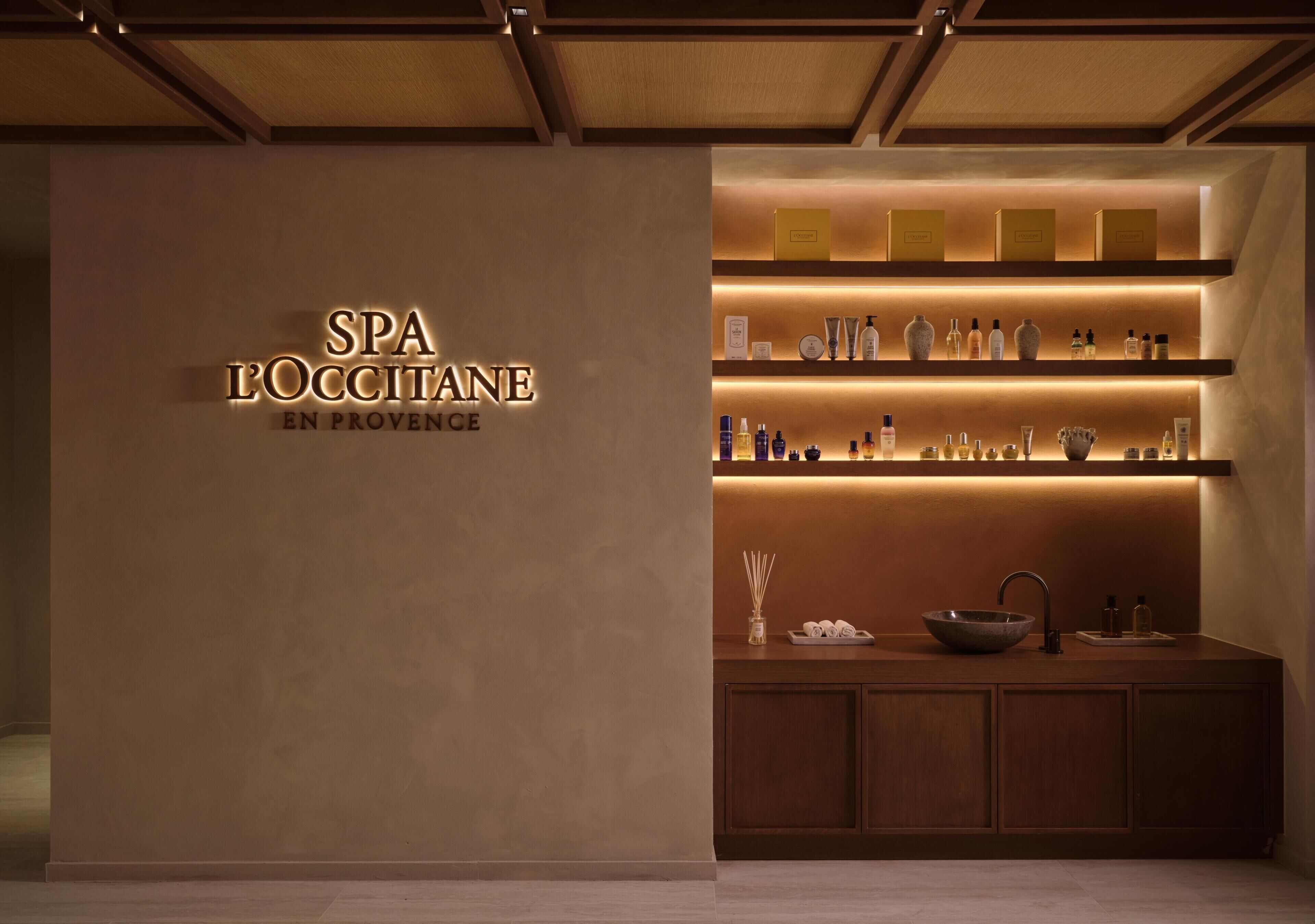 A spa reception area with a sign reading 'SPA L'OCCITANE EN PROVENCE,' shelves with various products, a sink, and a vase.