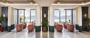 Una hall d'albergo con vista sulla spiaggia, con un'area salotto con pouf decorati e una pianta in vaso al centro.