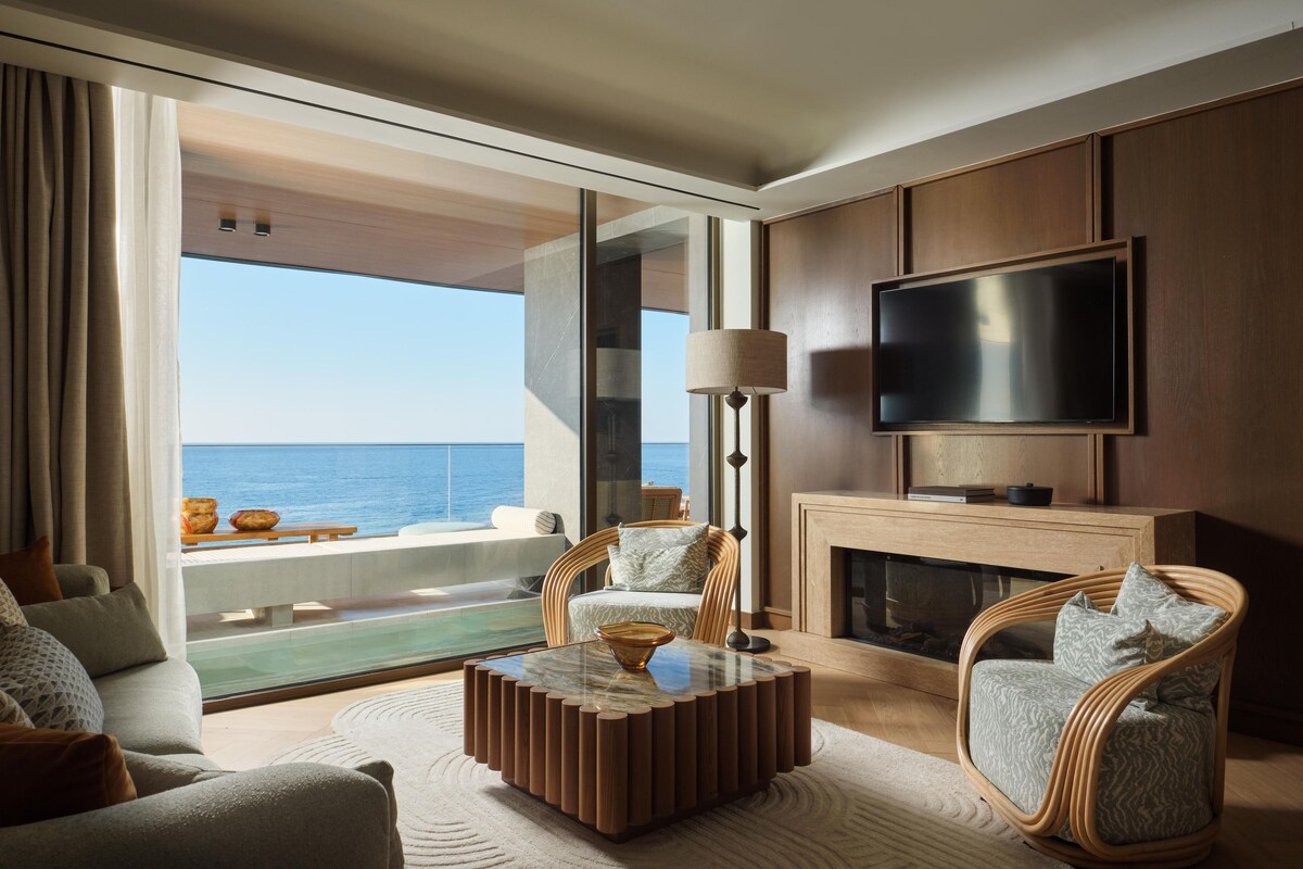 A modern living room with a large TV, comfortable seating, a fireplace, and a view of the ocean.
