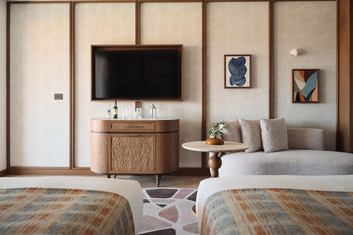 A hotel room with a flat-screen TV, a minibar, a sofa, two beds, and a patterned rug.