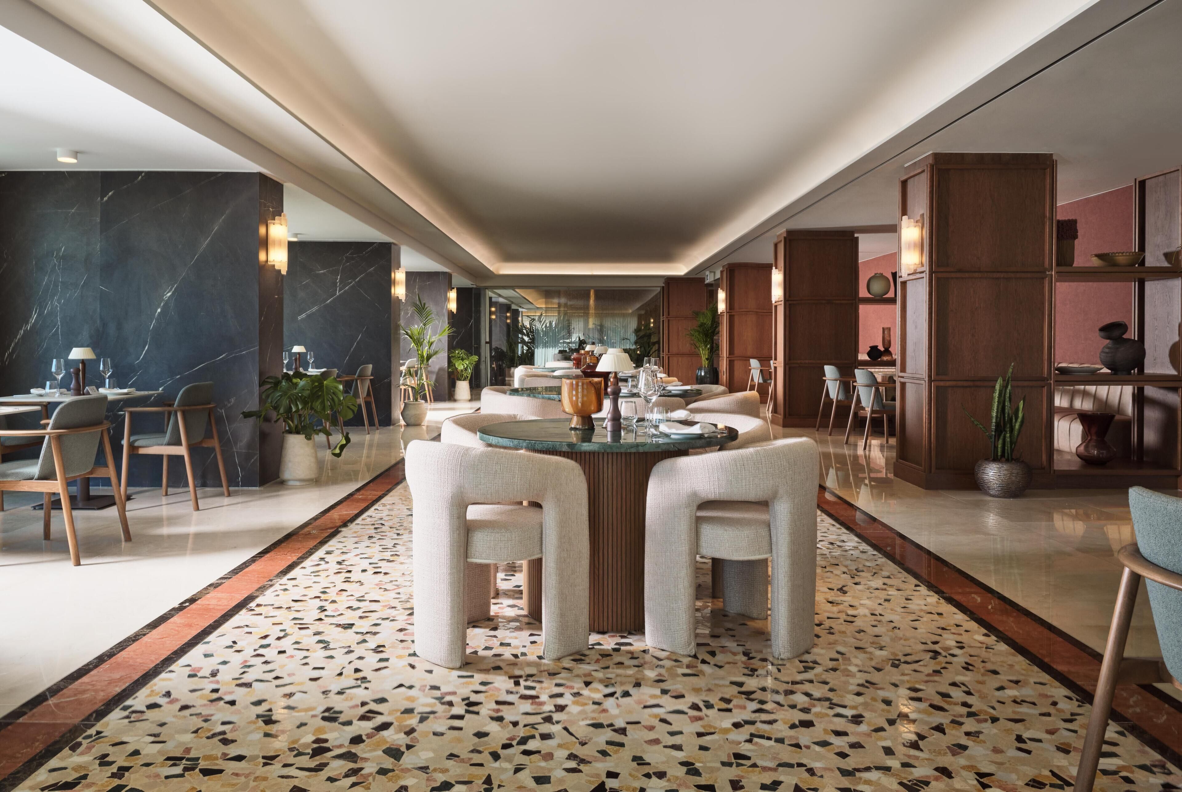 Un moderno vestíbulo de hotel con una mesa de comedor redonda, dos sillones blancos, un suelo estampado y detalles de mármol y madera.