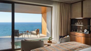 Habitación de hotel con un amplio ventanal corredero que da a un balcón y vistas al mar. Cuenta con una cama con colcha estampada, una mesita redonda con un florero y un rincón de descanso con una silla y una mesa.