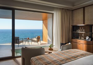 A hotel room with a large sliding glass door leading to a balcony with a view of the sea. There is a bed with a patterned bedspread, a small round table with a vase of flowers, and a seating area with a chair and a table. - Hilton Chania Old Town Resort & Spa (Chania)