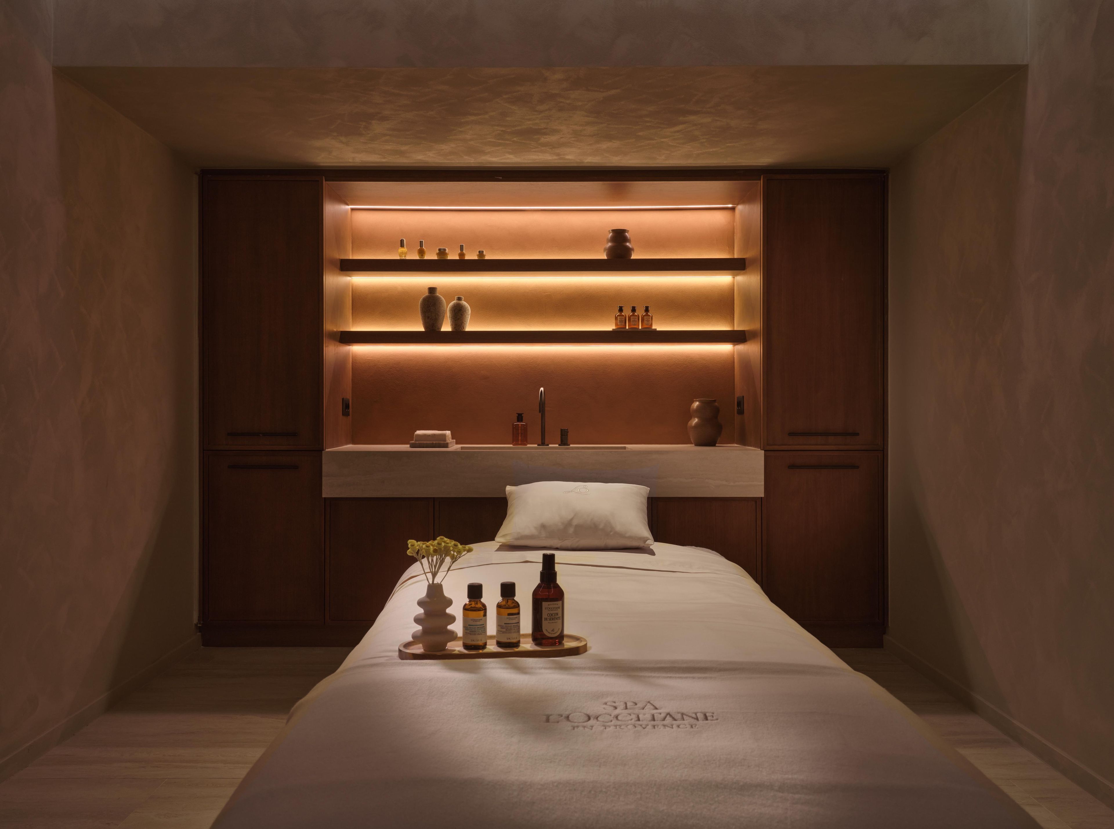 A hotel room with a bed, a bedside table with toiletries, a shelf with decorative items, and a wall-mounted shelf with bottles.