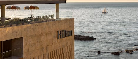 A hotel with a view of the sea, a sign reading 'Hilton,' outdoor umbrellas, and a boat in the distance.