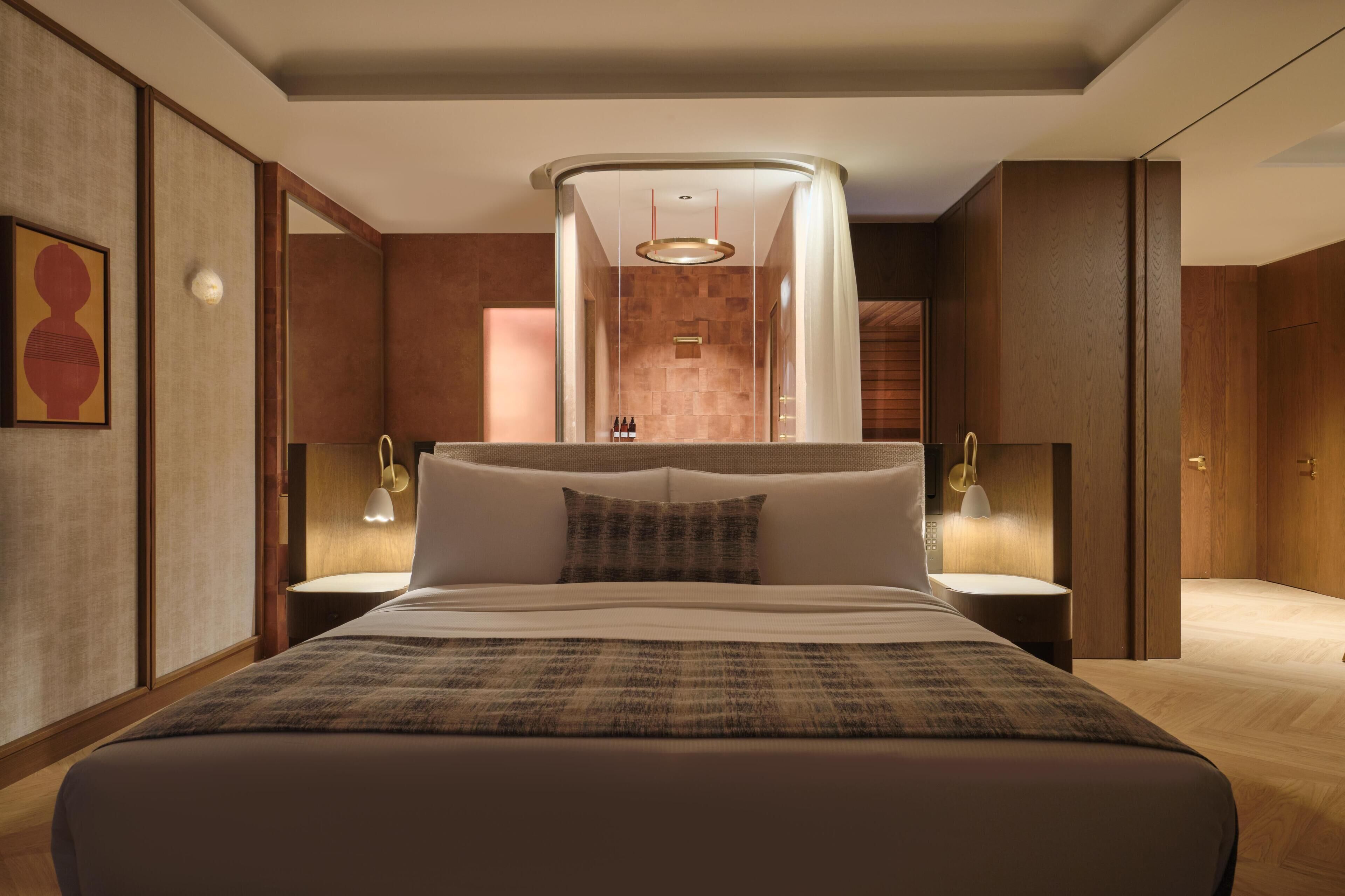 Ein modernes Hotelzimmer mit einem großen Bett, zwei Nachttischen, einer Wandleuchte und Blick auf den Flur.