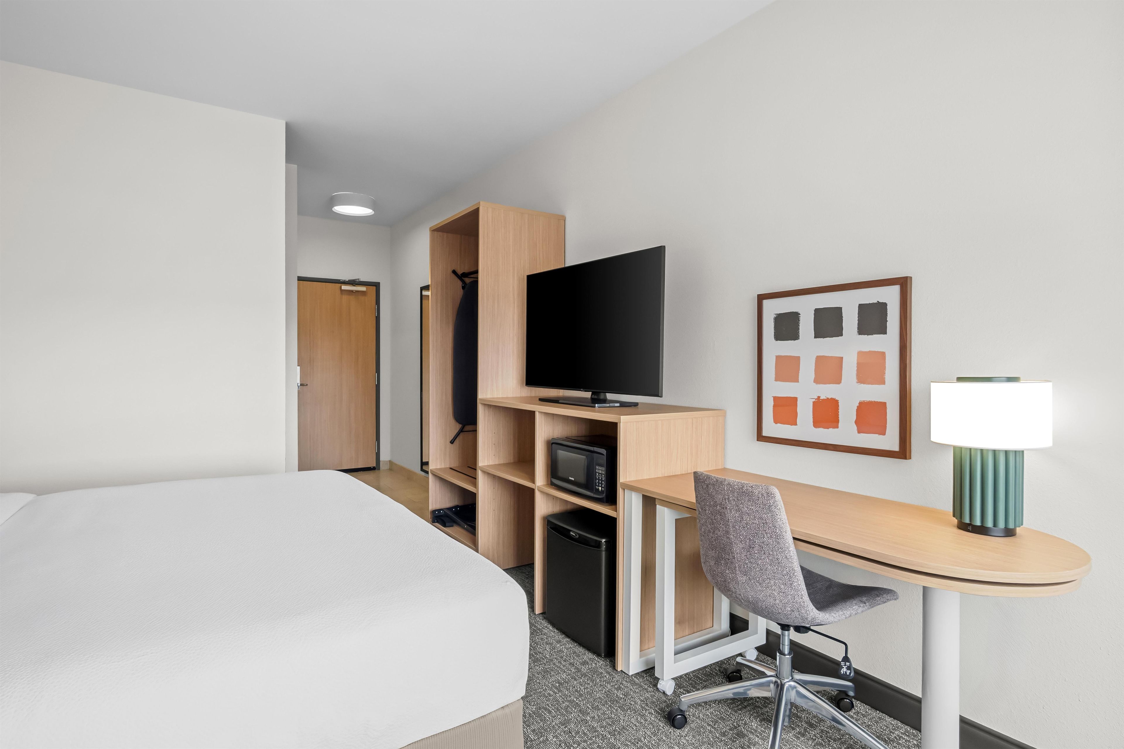 Habitación de hotel con una cama, un escritorio con silla, un televisor, un microondas y un cuadro en la pared.