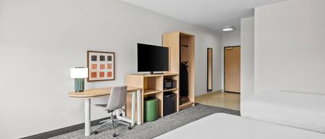 A hotel room with a bed, a desk with a chair, a television, a wardrobe, and a small trash bin.