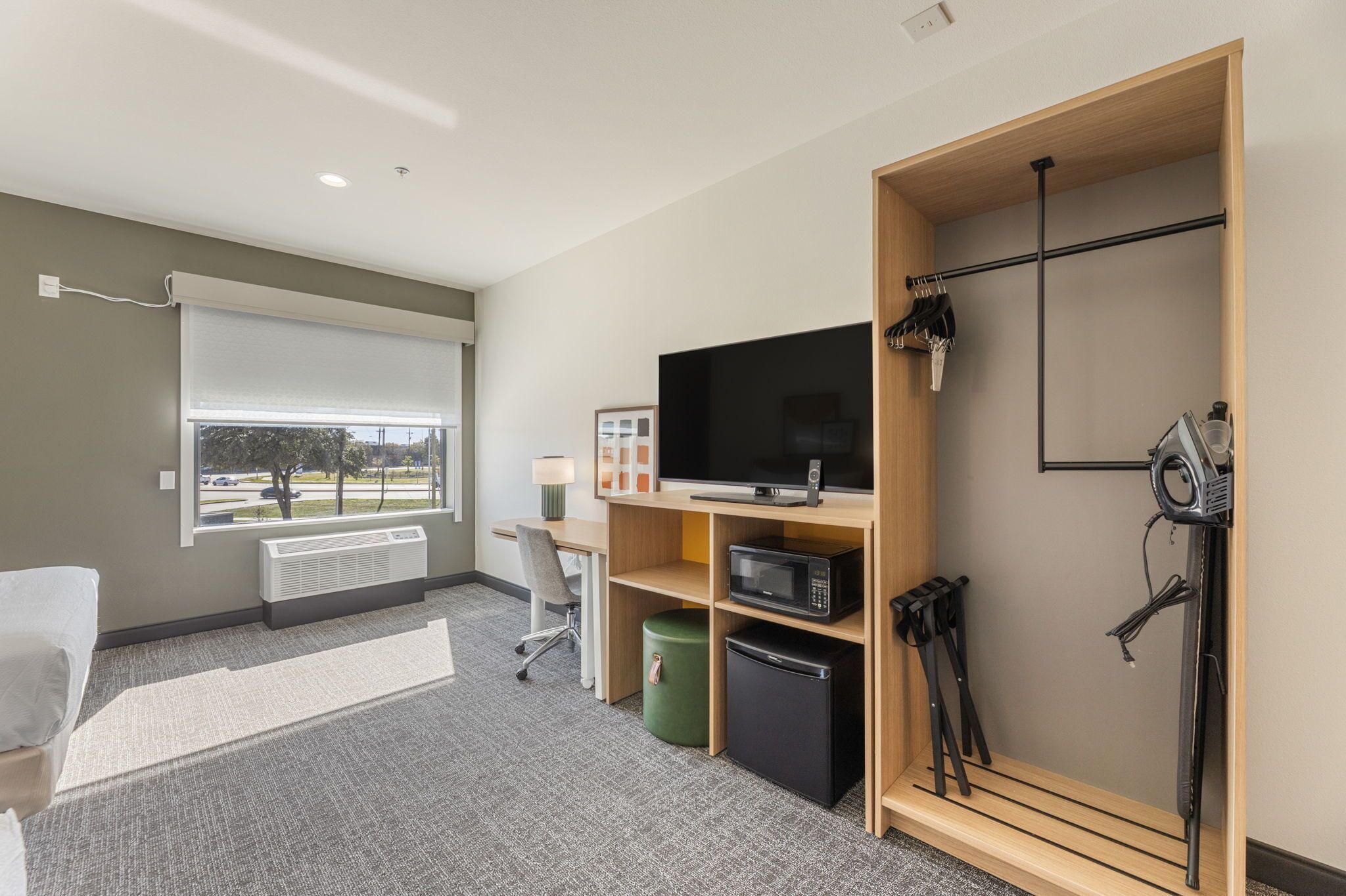 Una habitación de hotel moderna con un ventanal grande, un televisor de pantalla plana, un escritorio con una silla, un microondas, un refrigerador pequeño y un perchero.