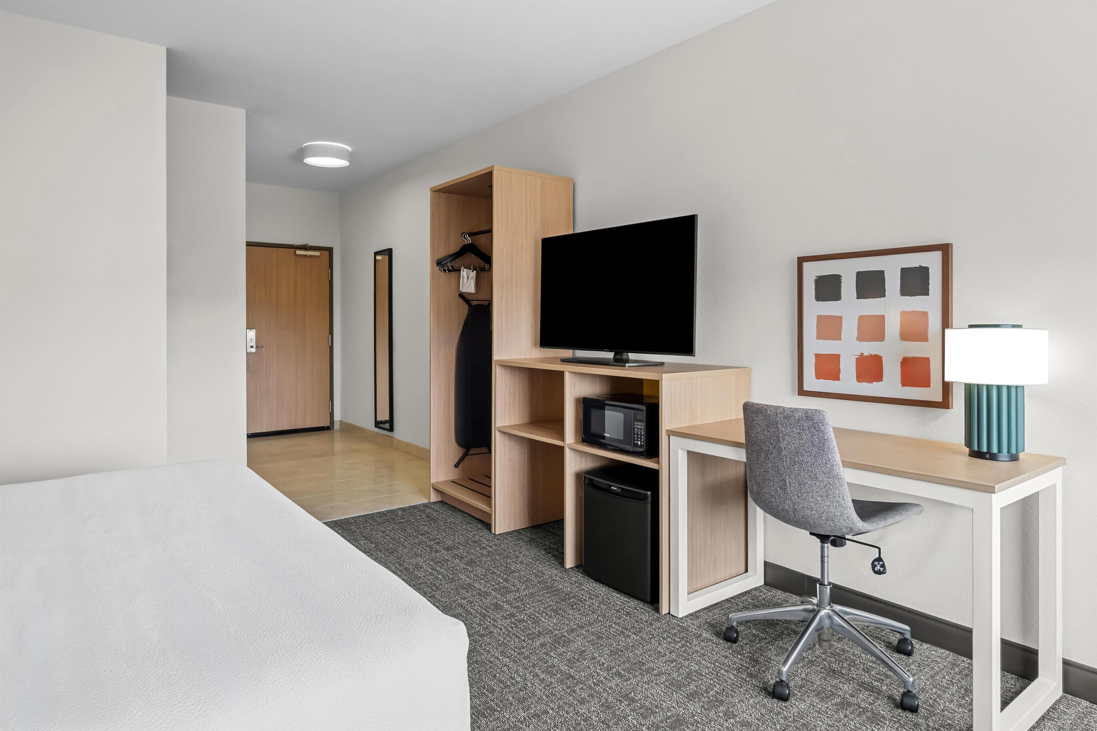 A compact hotel room with a bed, a desk with a computer, a television, a microwave, and a small refrigerator.