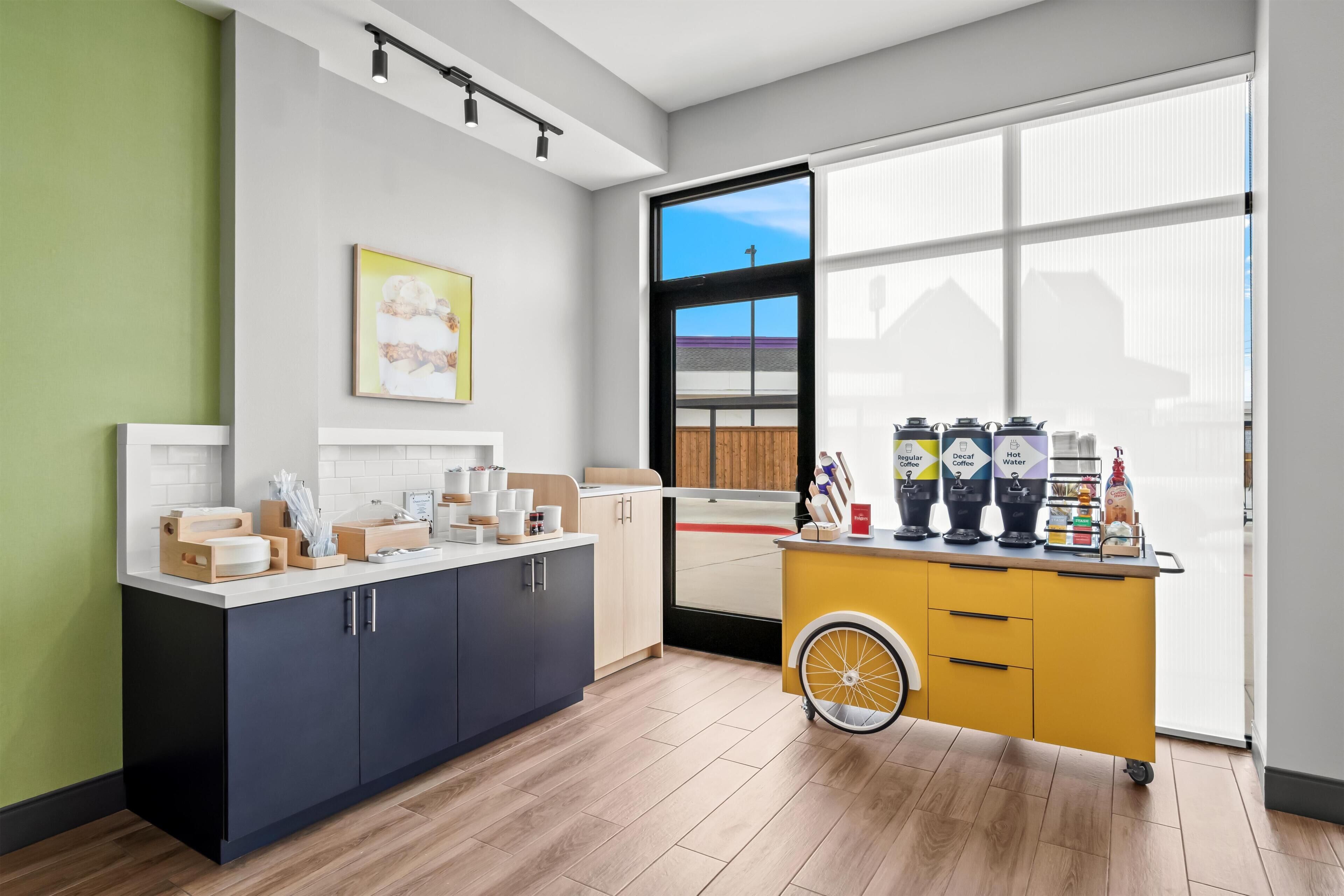 A modern kitchen with a large window, a yellow cart with coffee machines, and a green wall.