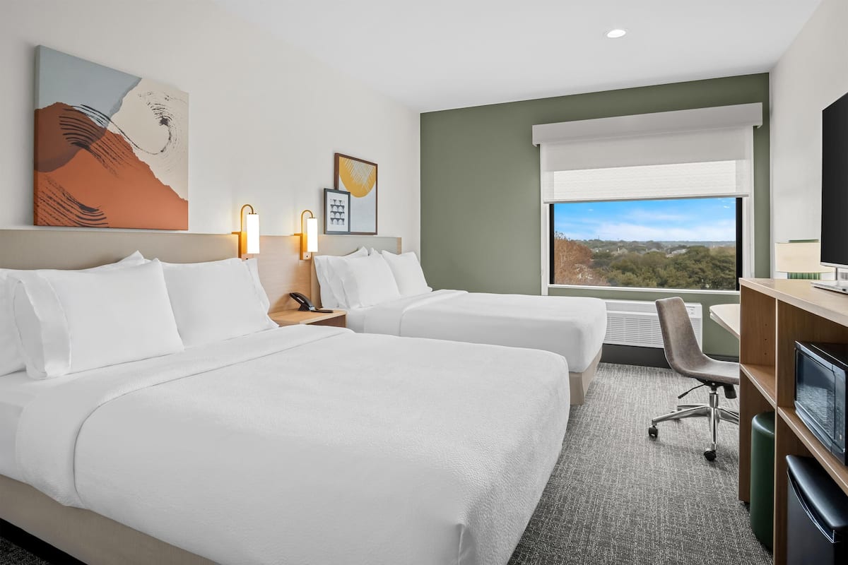 Ein Hotelzimmer mit zwei Betten, einem Schreibtisch, einem Fernseher und einem großen Fenster mit Blick auf die Stadt.