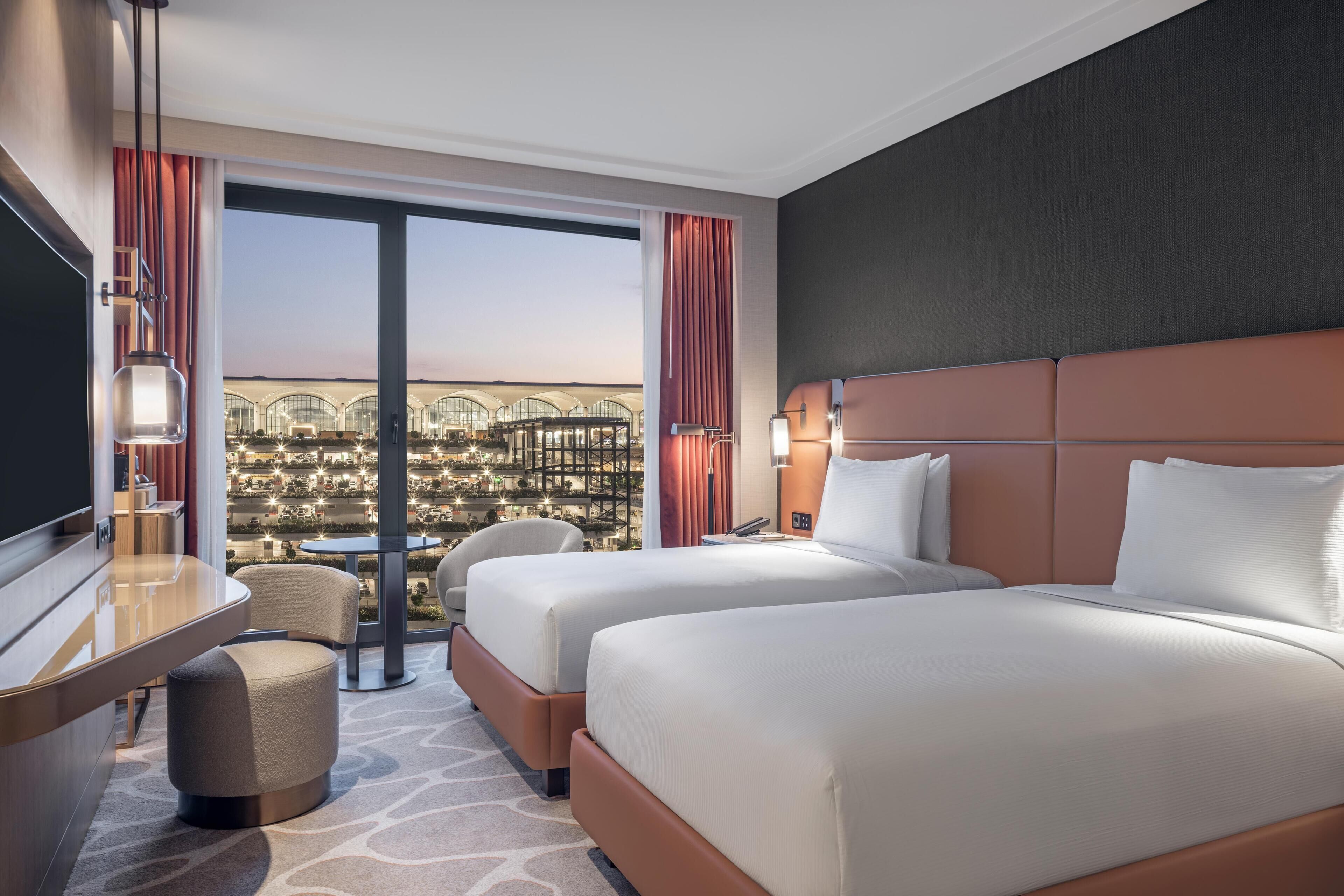 Quarto de hotel com duas camas, uma escrivaninha, uma cadeira e vista para a cidade.