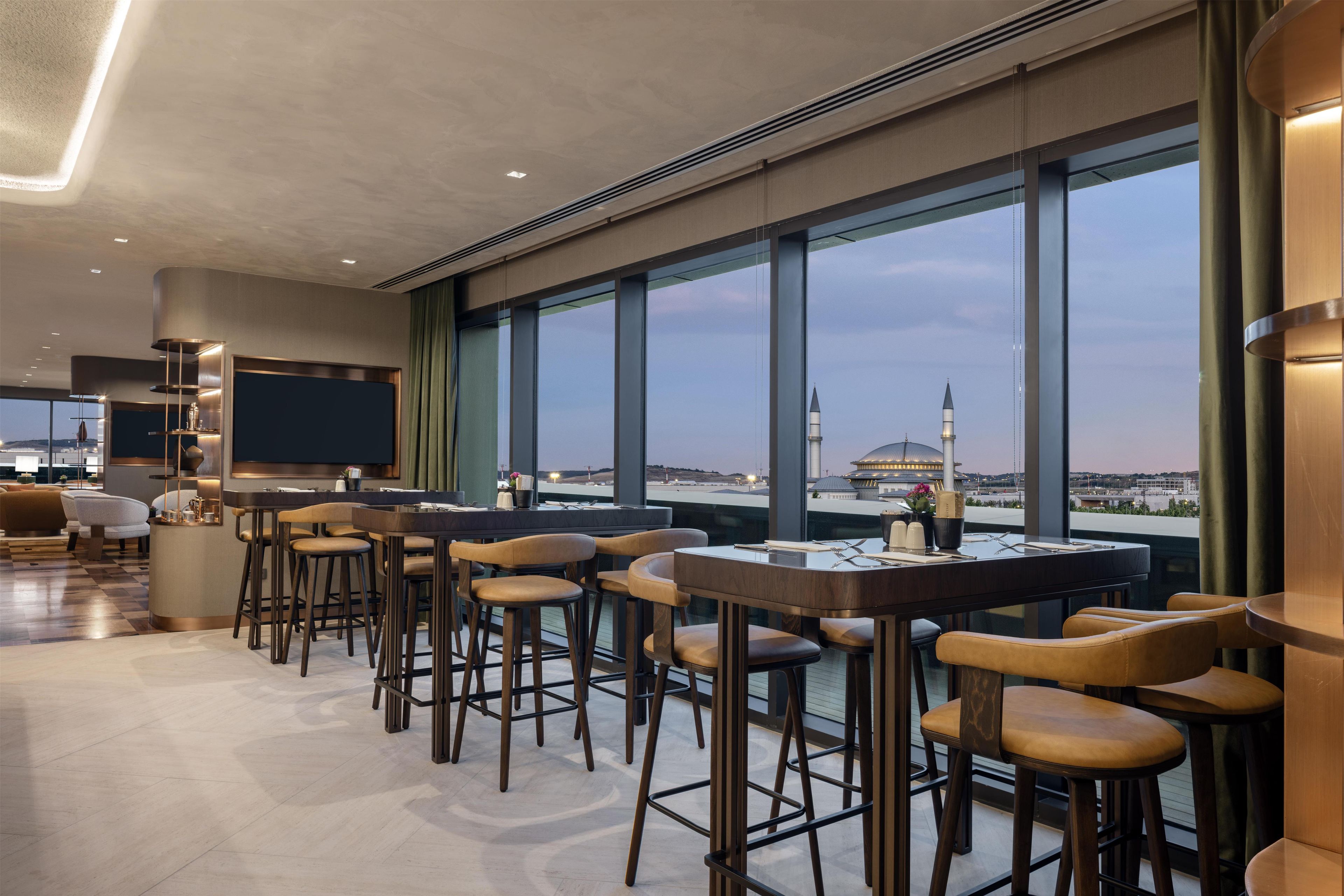 Zona de bar moderna com mesas e banquetas altas, TV de tela plana e grandes janelas com vista para a cidade.