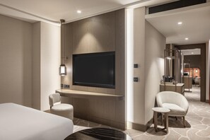 Quarto de hotel moderno com uma televisão de tela plana grande fixada em um painel de madeira, uma pequena mesa redonda, duas poltronas estofadas e uma cama com cabeceira branca.