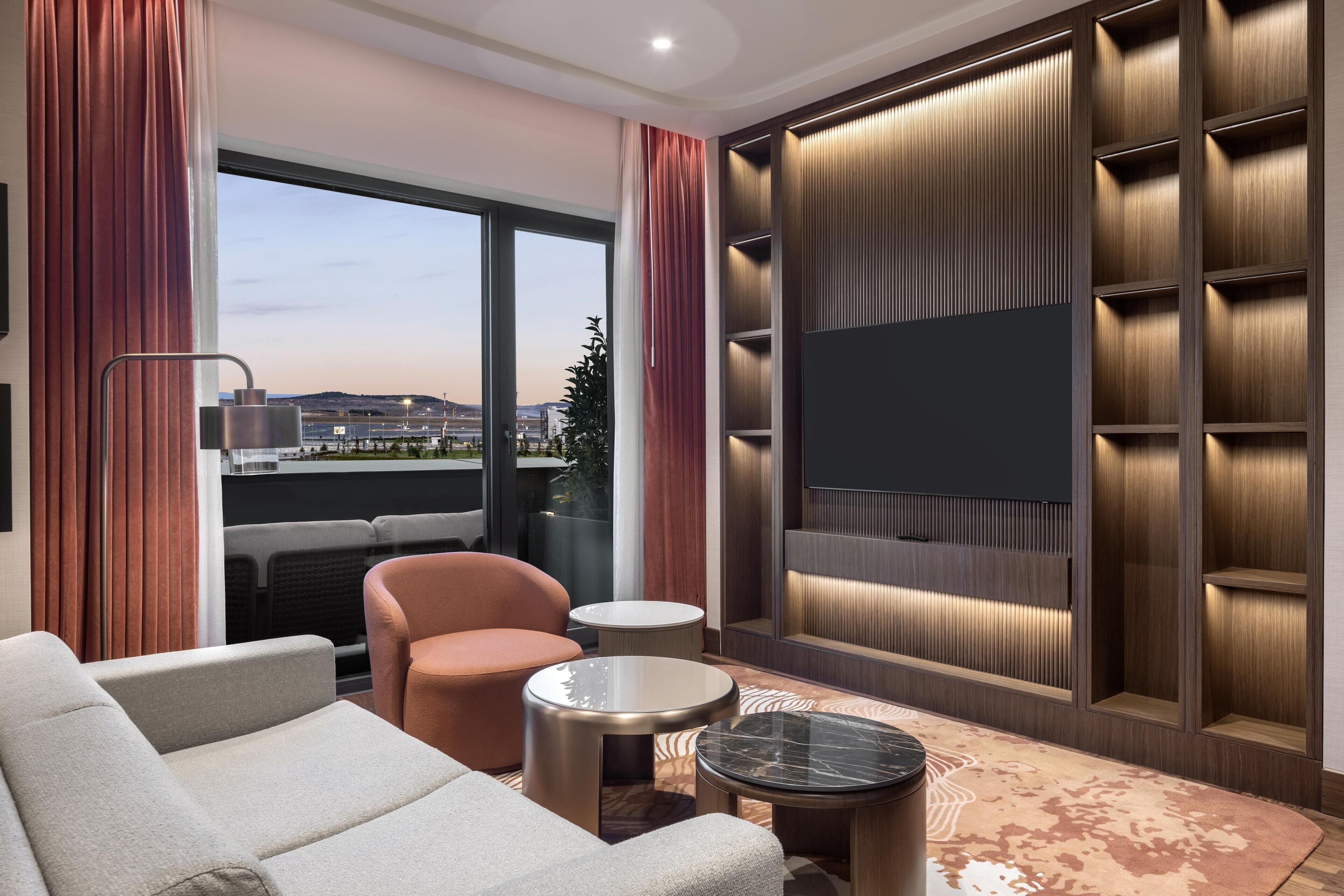 A modern hotel room with a large window, a sofa, a chair, a small table, and a TV mounted on a wooden panel wall.