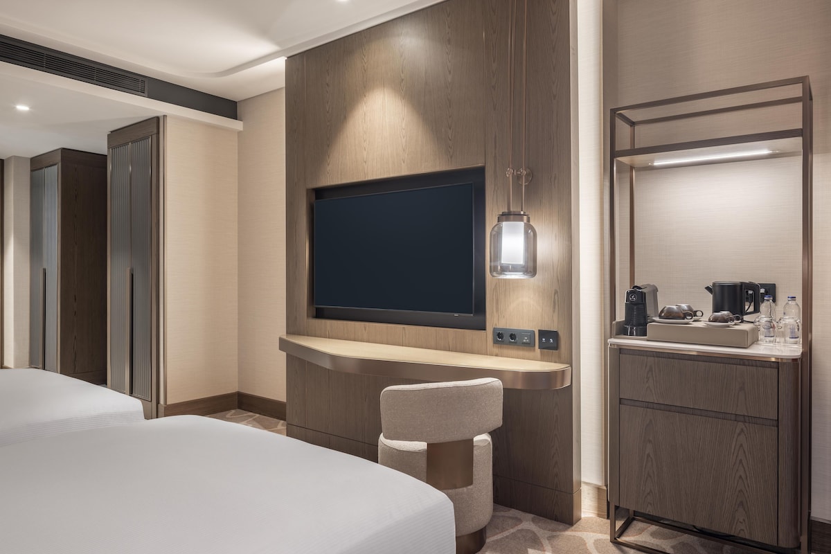 A hotel room with a flat-screen TV mounted on a wooden panel, a small seating area with a cushioned chair, a bedside table with a coffee maker, and a minibar.