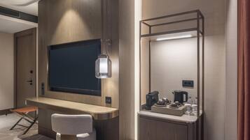 Ein Hotelzimmer mit Flachbildfernseher an einer Holzwand, einem Klappschreibtisch mit Stuhl, einem kleinen Tisch mit Kaffeemaschine und Tassen sowie einem Spiegel mit Beleuchtung.