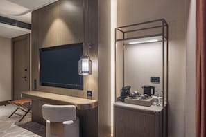 Quarto de hotel com televisão de tela plana fixada em parede de madeira, uma escrivaninha dobrável com cadeira, uma mesinha com cafeteira e xícaras, e um espelho com iluminação.