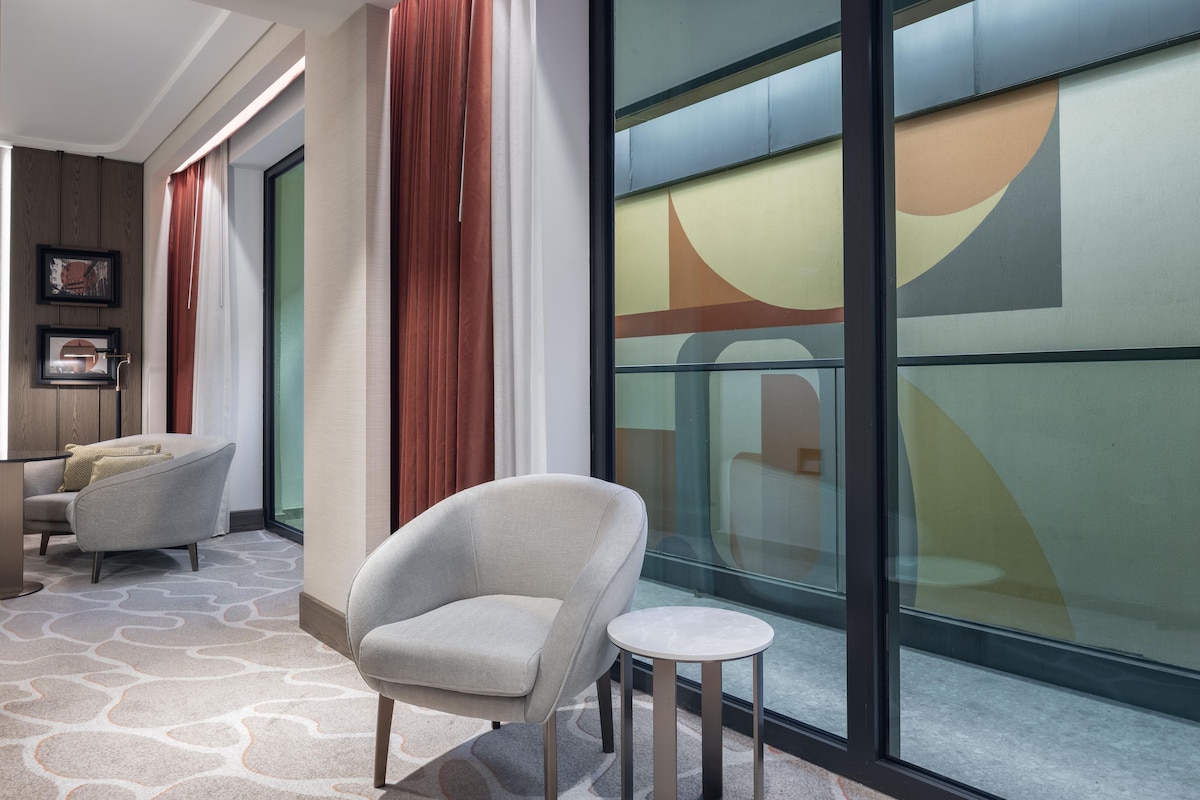 A modern interior with a grey armchair, a small round table, and a large window with abstract wall art.
