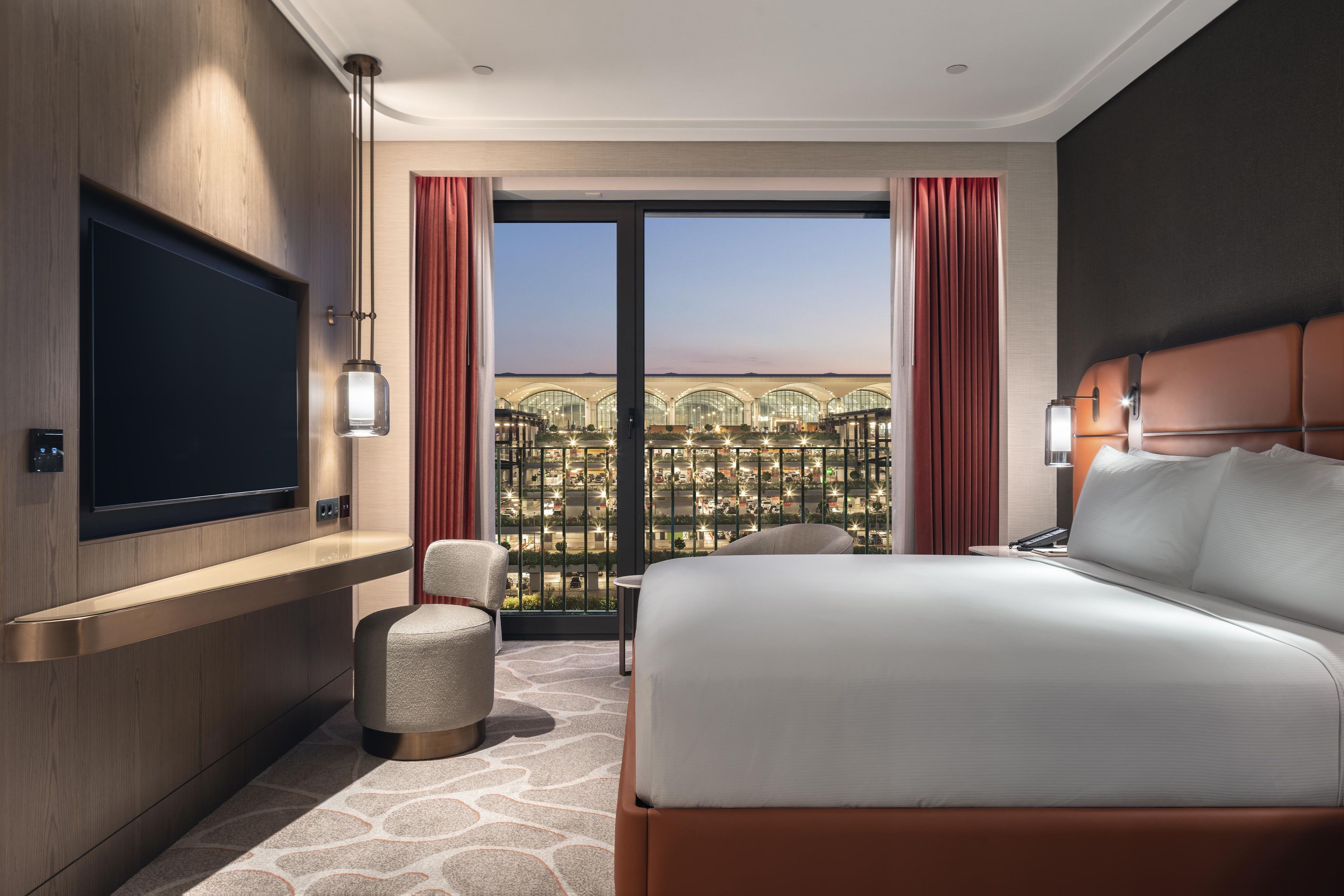 A hotel room with a large bed, a flat-screen TV, a seating area with chairs, and a view of a building with multiple levels.