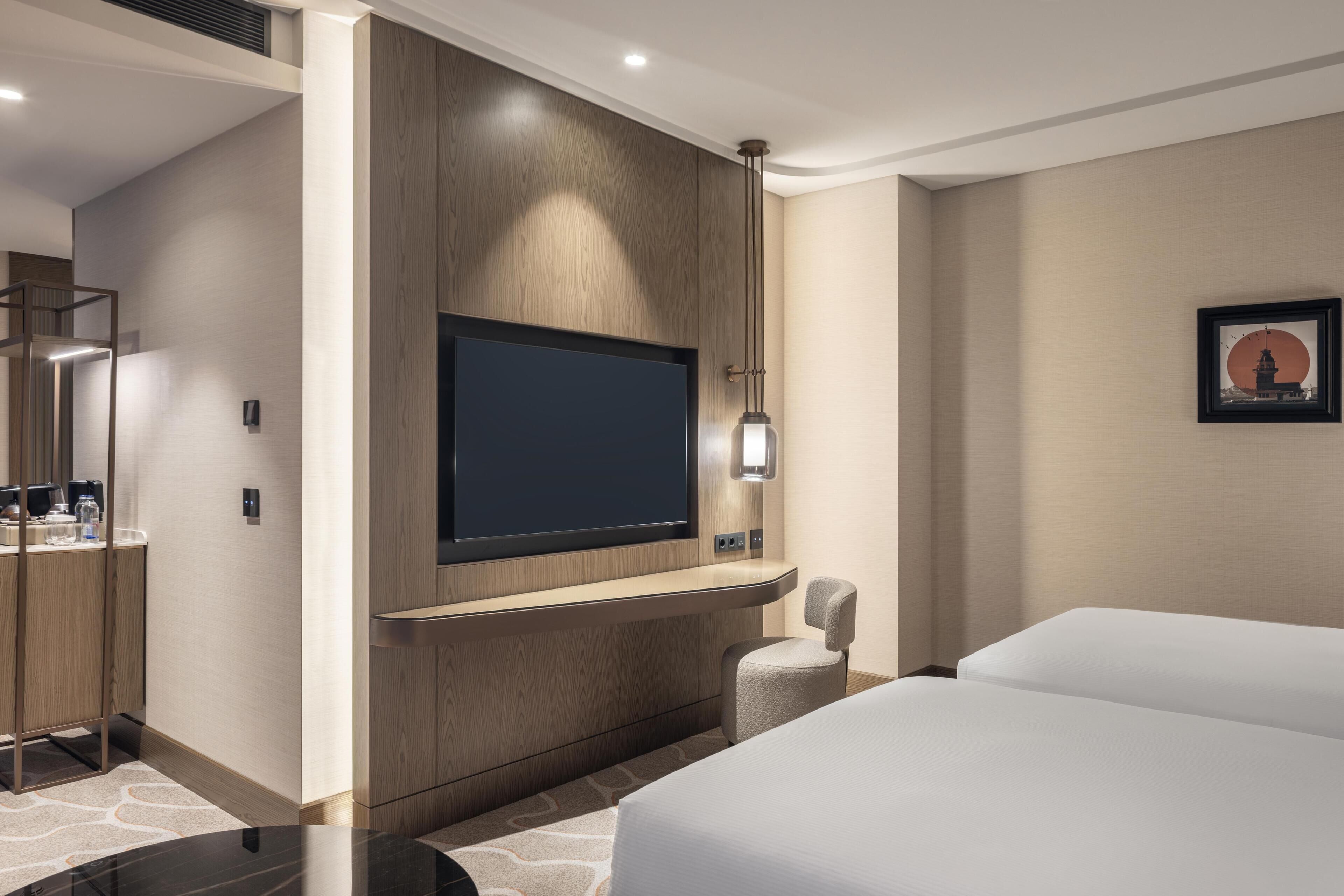 A hotel room with a bed, a flat-screen TV mounted on a wooden panel, a small table, and a chair.