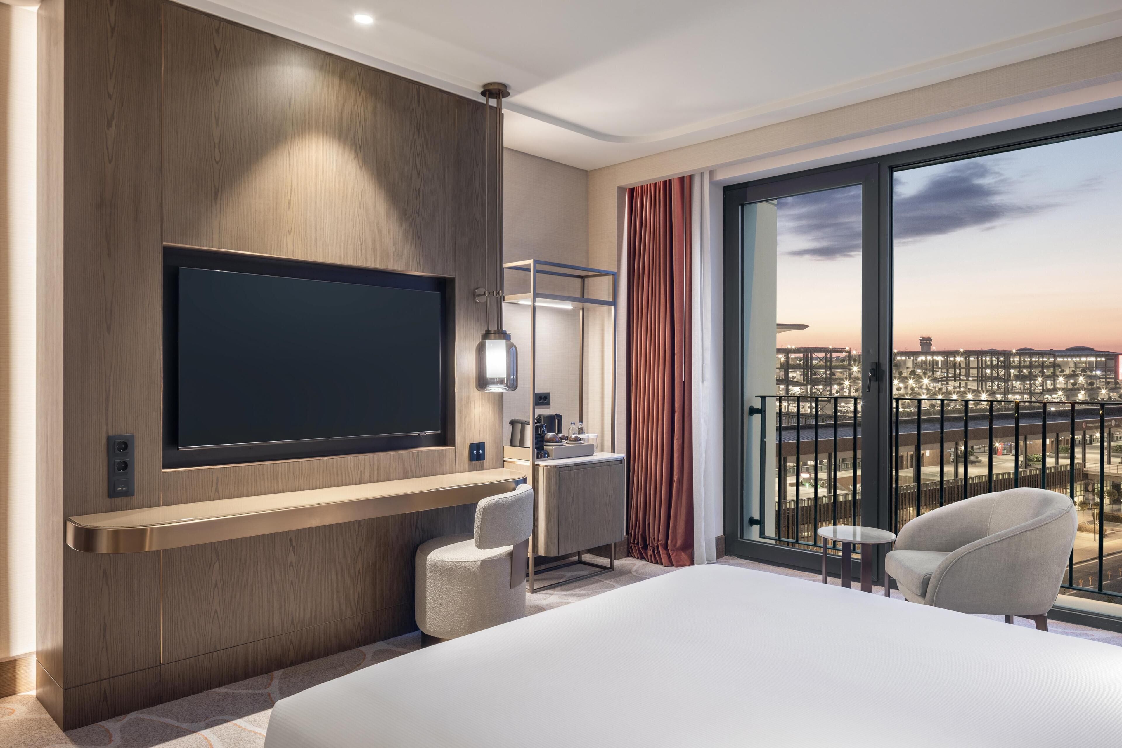 A hotel room with a large bed, a flat-screen TV mounted on the wall, a desk with a chair, a small round table, and a balcony with a view of the city.